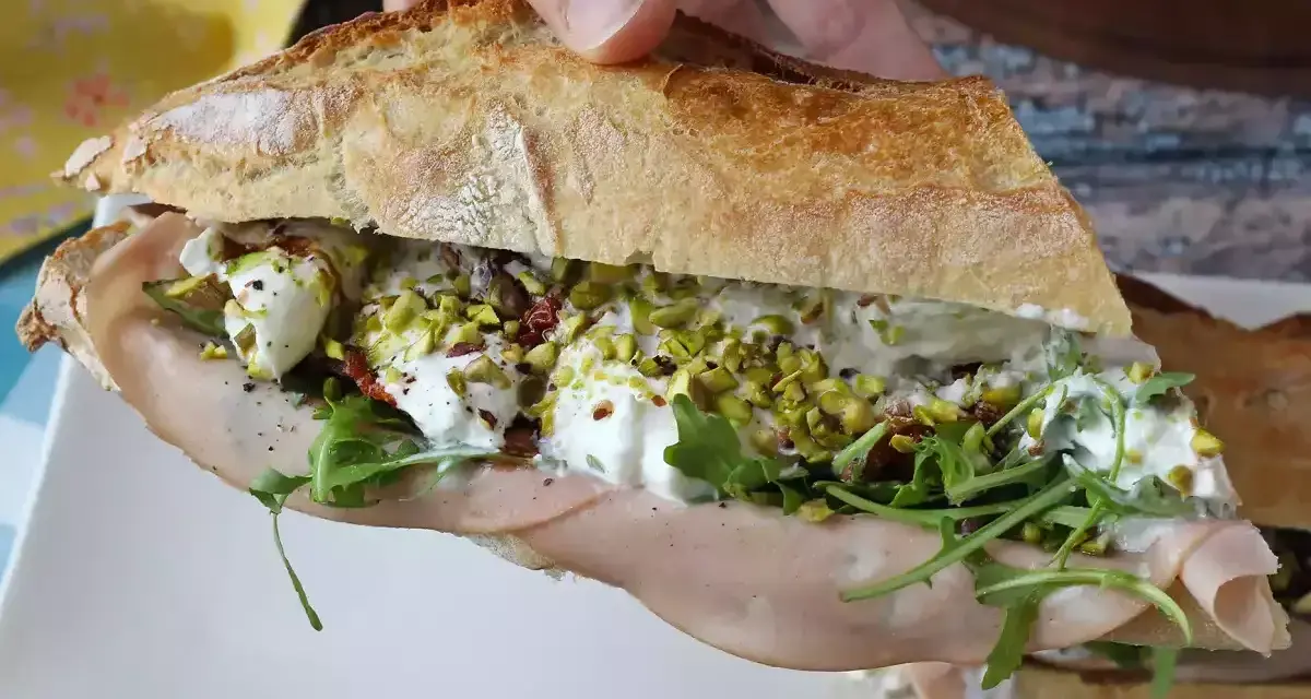 Bocadillo de pan rústico relleno de mortadela, burrata cremosa, rúcula, pistachos y tomates secos.