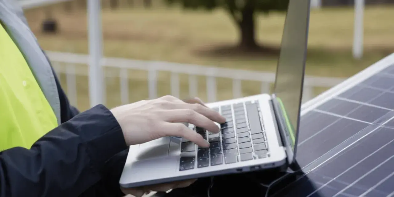 Praca nad projektem fotowoltaiki: ręce na klawiaturze laptopa, obok panel słoneczny.