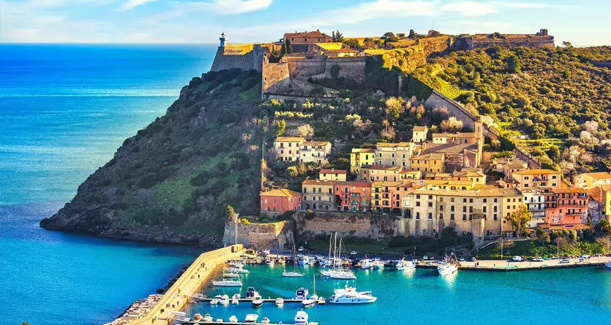Incantevoli toscana paesaggi: un borgo colorato su una collina fortificata domina un porto con barche, baciato dal sole.
