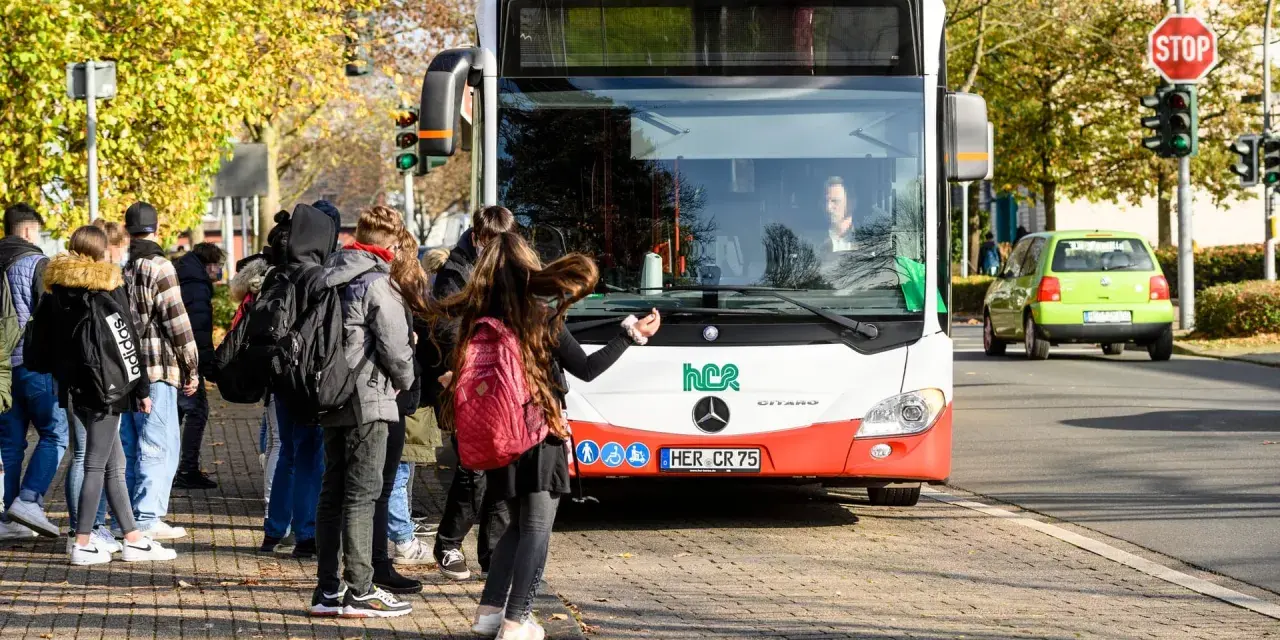 Ein Schulbus mit Warnblinker hält an einer Haltestelle. Schüler warten auf dem Gehweg, um einzusteigen.
