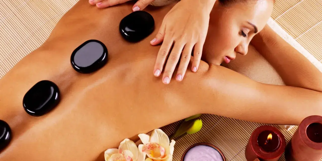 A woman enjoys the benefits of hot stone massage, with smooth black stones placed on her back. Orchids and candles add to the serene spa atmosphere.