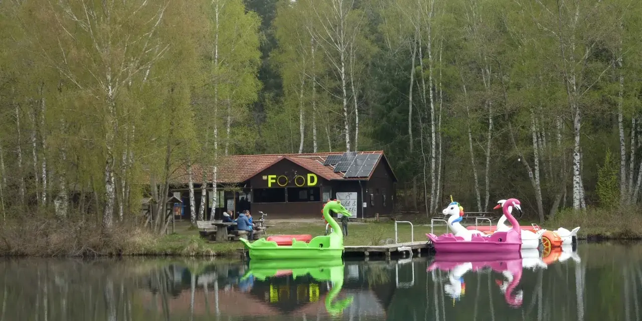 Bunte Tretboote in Form von Schwänen und Einhörnern liegen am Ufer des Bruchsee Duingen. Ein Gebäude mit der Aufschrift "FOOD" und Solaranlagen steht im Hintergrund.