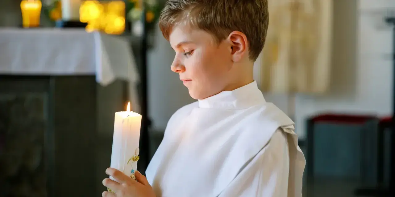 Chłopiec w bieli trzyma zapaloną świecę podczas uroczystości chrztu świętego.