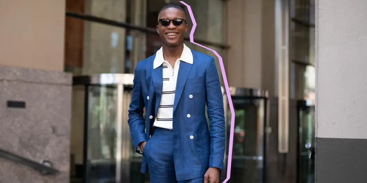 Hombre sonriente con traje azul y camiseta blanca a rayas, gafas de sol, posando con la mano en el bolsillo.