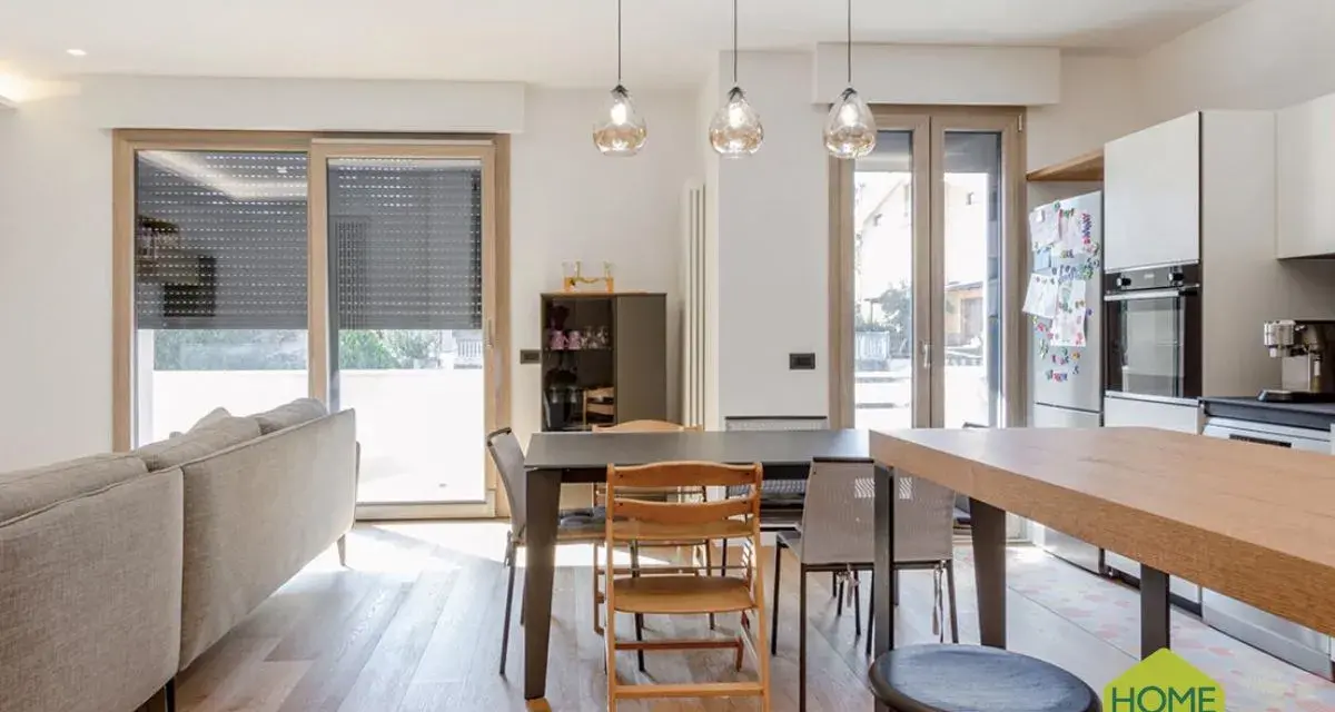 Un soggiorno luminoso con un divano, tavolo da pranzo e cucina a vista. Un moderno sopraluce illumina l'ambiente.