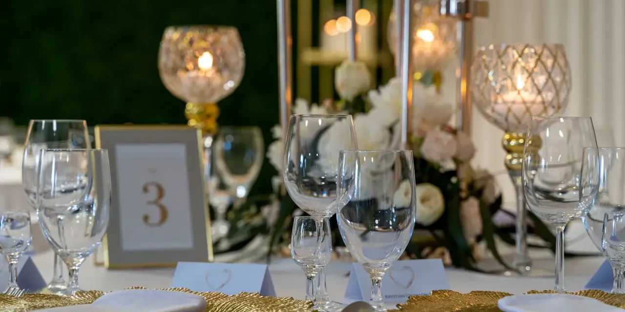 Elegancko nakryty stół weselny z kieliszkami, złotymi talerzami i świecami. Czy warto personalizować każdy element wesela od serwetek po etykiety? Tak!