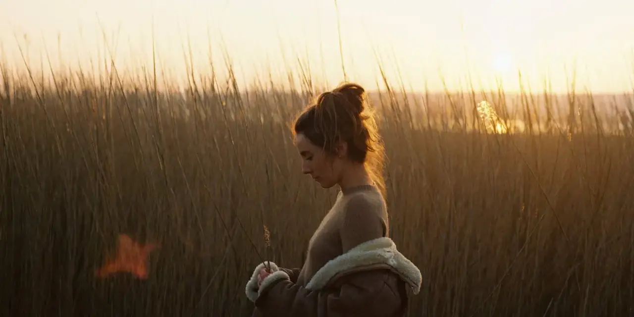 Une jeune femme, le regard baissé, se tient dans les hautes herbes au crépuscule. La lumière dorée du soleil couchant illumine ses cheveux et le paysage.