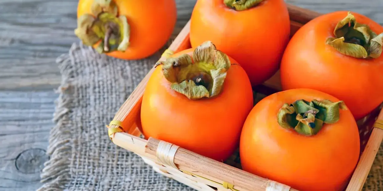 Frische, orangefarbene Kaki-Früchte liegen in einem kleinen Korb auf grobem Leinen. Kaki gesund und lecker.