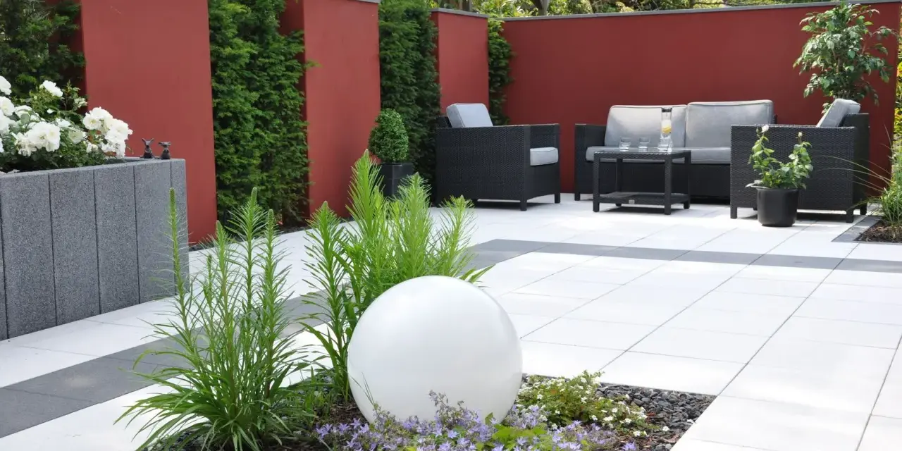 Moderner Garten ohne Rasen mit roten Mauern, weißen Terrassenplatten, dunklen Lounge-Möbeln und einer leuchtenden Kugel.