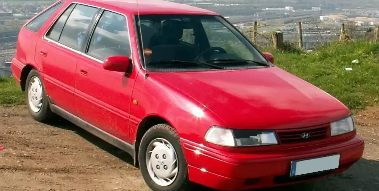 Czerwony Hyundai Pony z lat 90. stoi na polnej drodze, z widokiem na dolinę i miasto w tle.