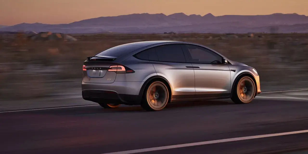 Srebrny Tesla X pędzi przez pustynny krajobraz o zachodzie słońca. Zastanawiasz się nad jego ceną?