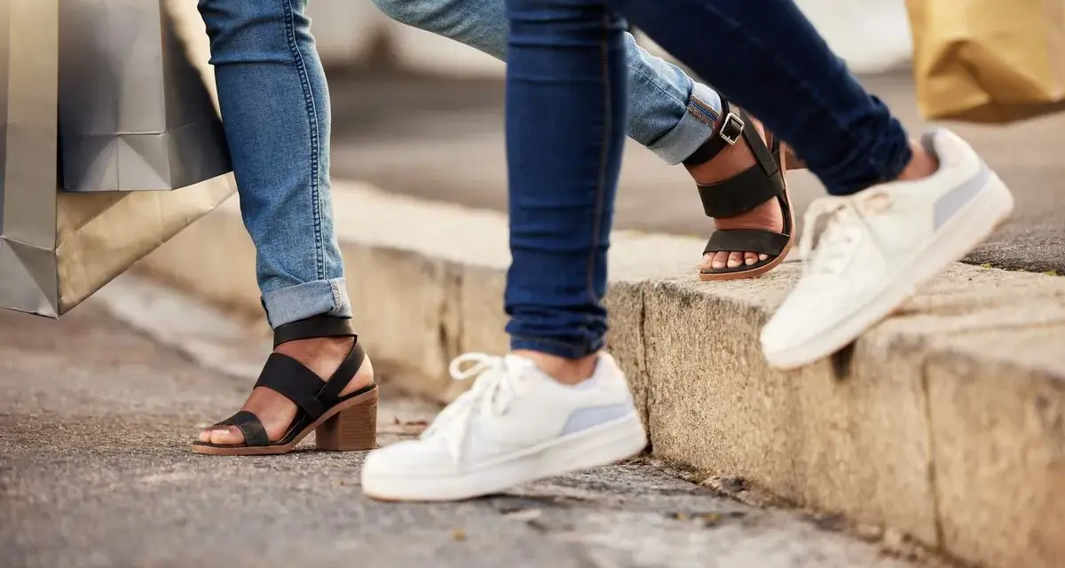 Dwie osoby w jeansach i modnych butach, z torbami na zakupy w rękach, idą chodnikiem. Jedna ma czarne sandały na obcasie, druga białe sneakersy.