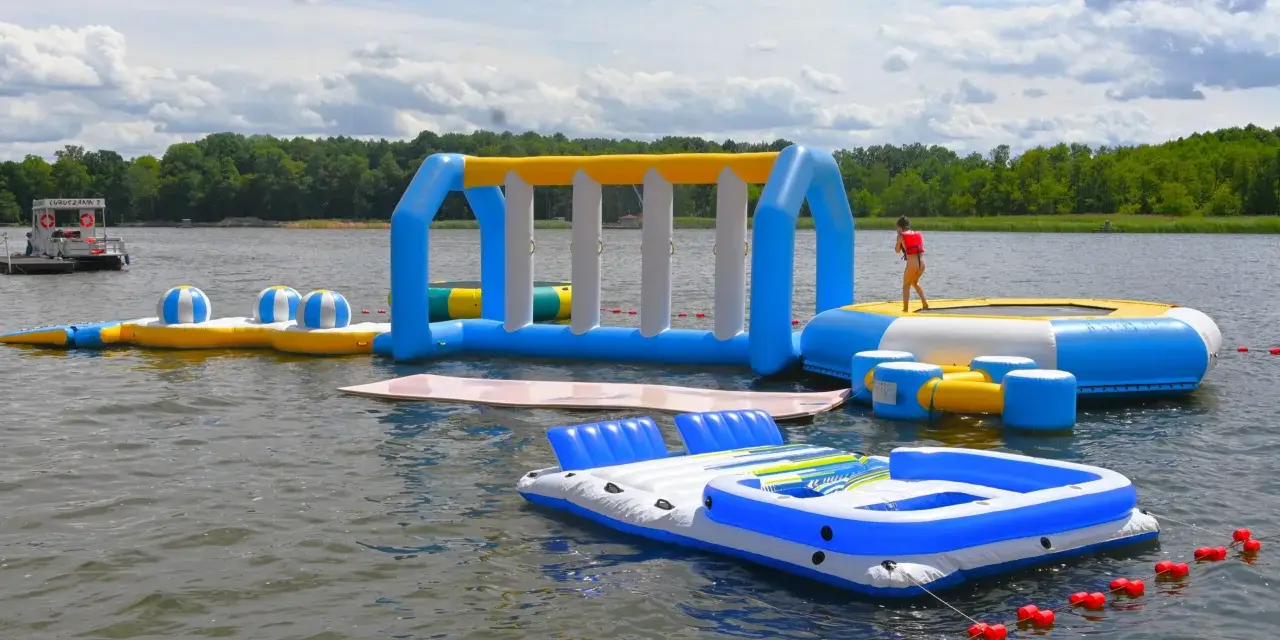 Kolorowe dmuchane atrakcje dla dzieci na jeziorze: zjeżdżalnia, trampolina i dmuchane wyspy. W tle las i łódka.