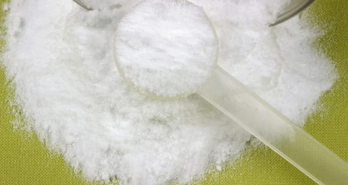 Biały proszek, prawdopodobnie l-glutamina, rozsypany na zielonym tle, z plastikową miarką.