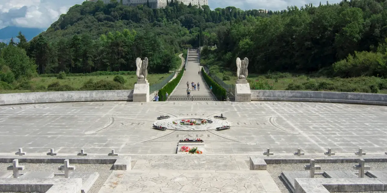Cmentarz żołnierzy na wzgórzu, z widocznym klasztorem na szczycie. Droga prowadzi do niego przez monumentalny plac z krzyżami i pomnikiem. To miejsce pamięci o bitwie pod **Cassino - trzeci / rzeka**.