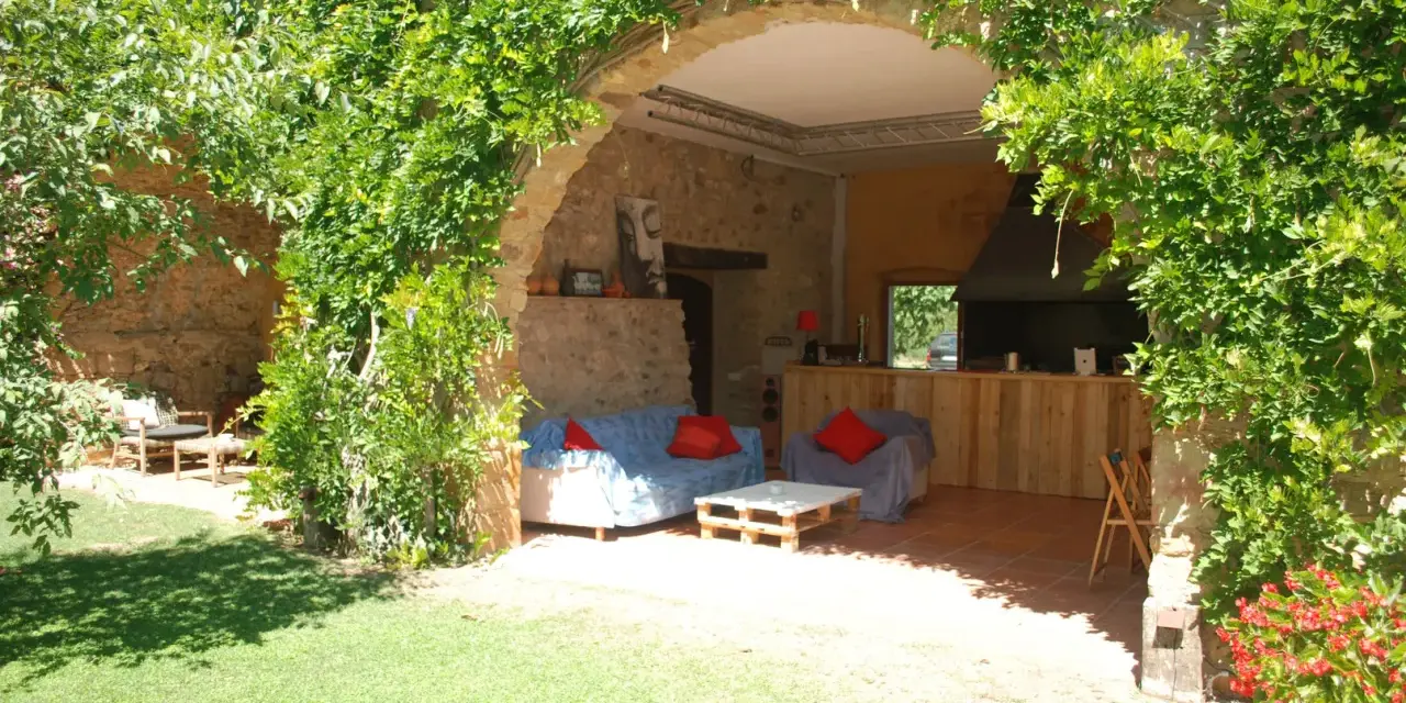 Un jardín con una madreselva planta cubriendo un arco de piedra. Dentro, sofás y una barra de madera.