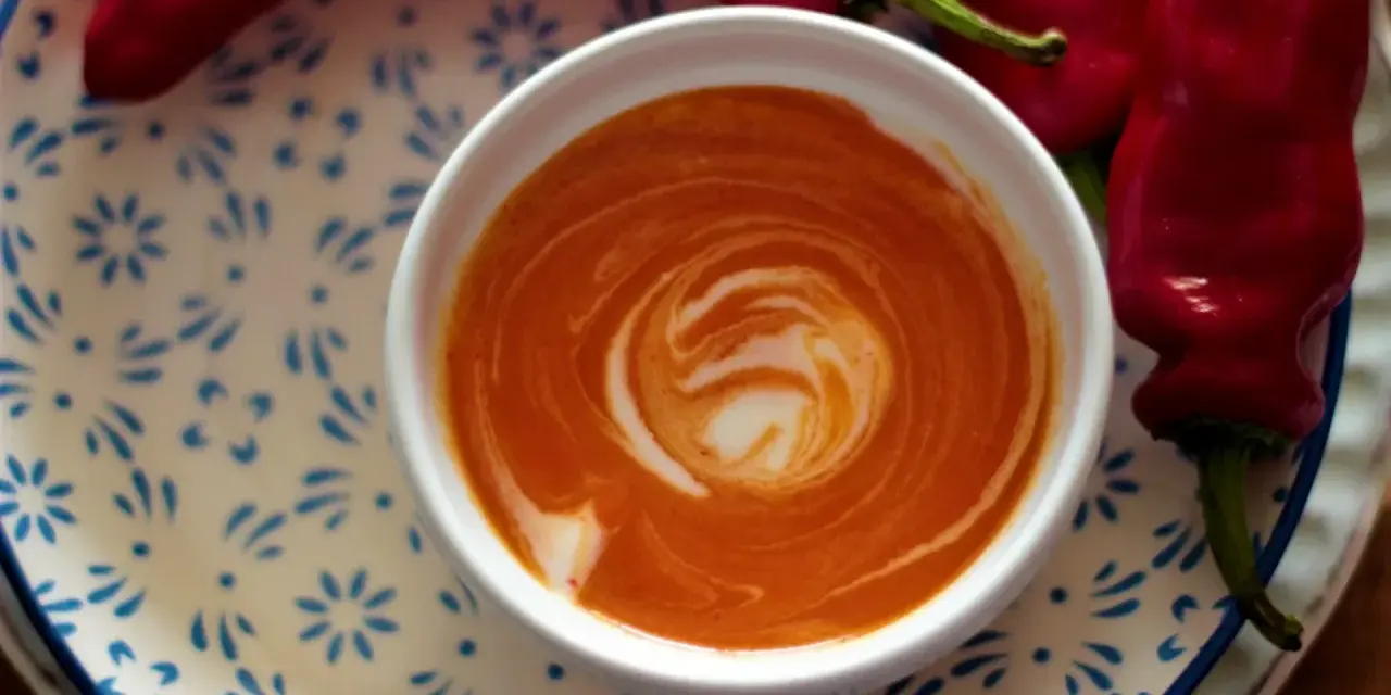 Crema di peperoni vellutata, con un vortice di panna, accanto a peperoni rossi freschi su un piatto decorato.