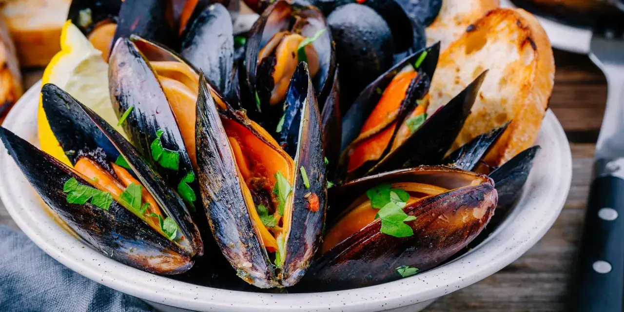 Mejillones en salsa, servidos en un cuenco blanco con perejil fresco, rodajas de limón y pan tostado.