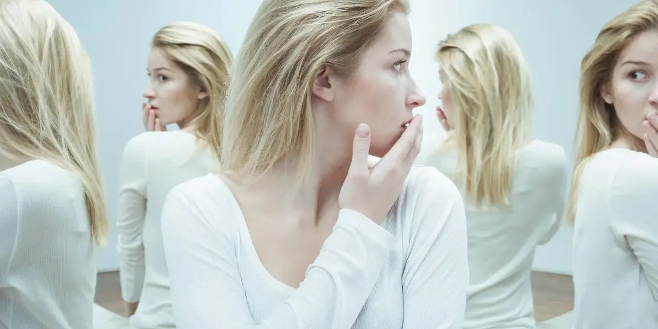 Kobieta z blond włosami w białej bluzce, otoczona swoimi odbiciami w lustrze.