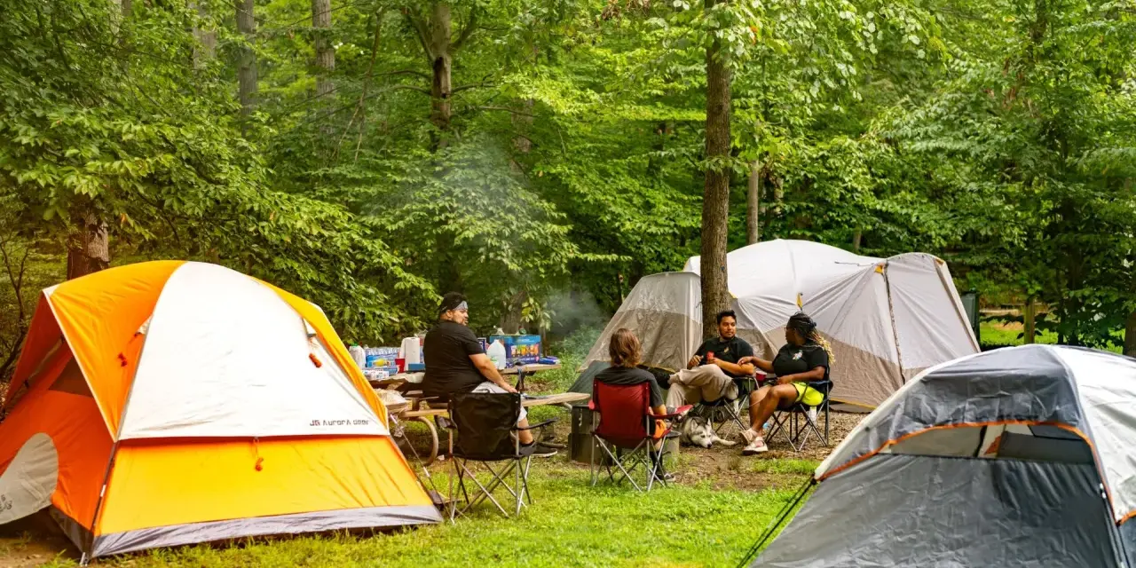 Grupa przyjaciół odpoczywa na polu namiotowym Mazury. Wokół rozstawione są namioty, a na stole piknikowym stoją napoje i przekąski.