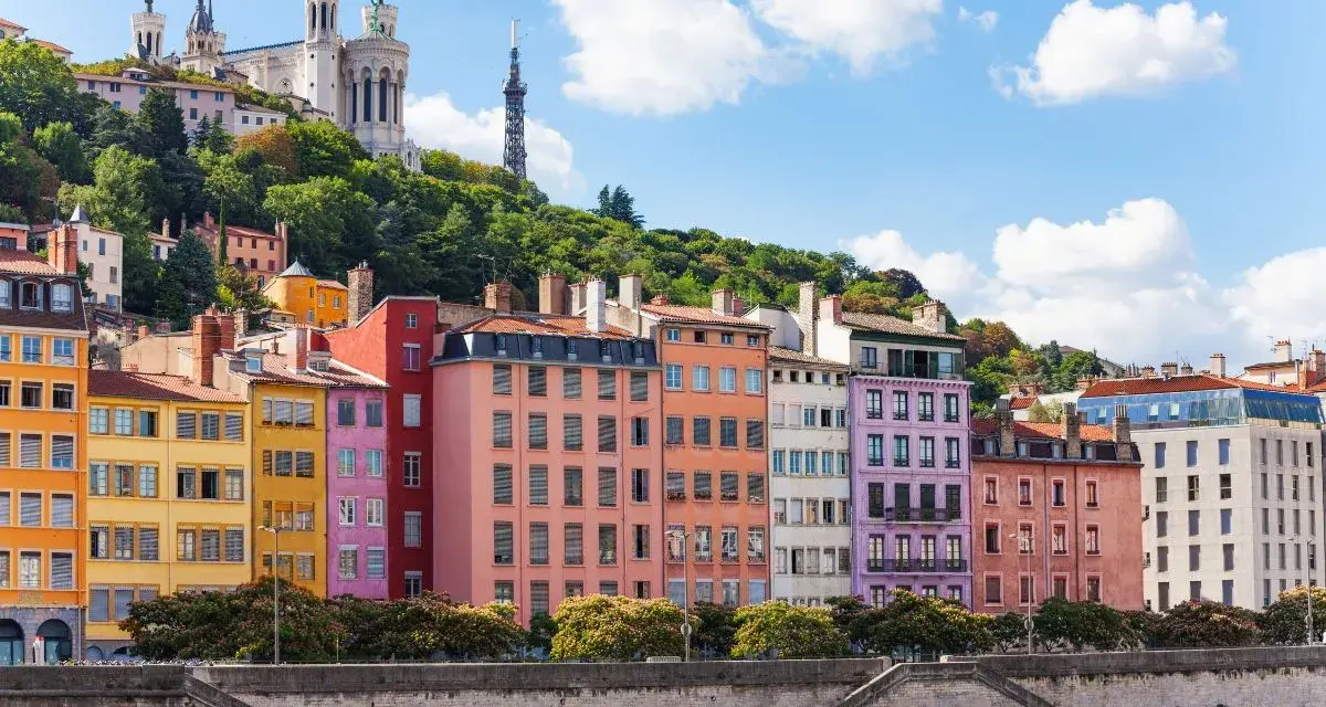 Colorful buildings line the river in Lyon, France, with a basilica on the hill above. A perfect scene for Lyon travel.