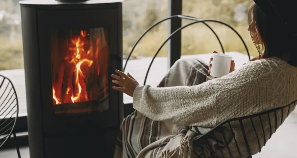 Donna avvolta in una coperta, con una tazza in mano, si scalda vicino a una stufa a legna, un modo accogliente per riscaldare la casa senza termosifoni.