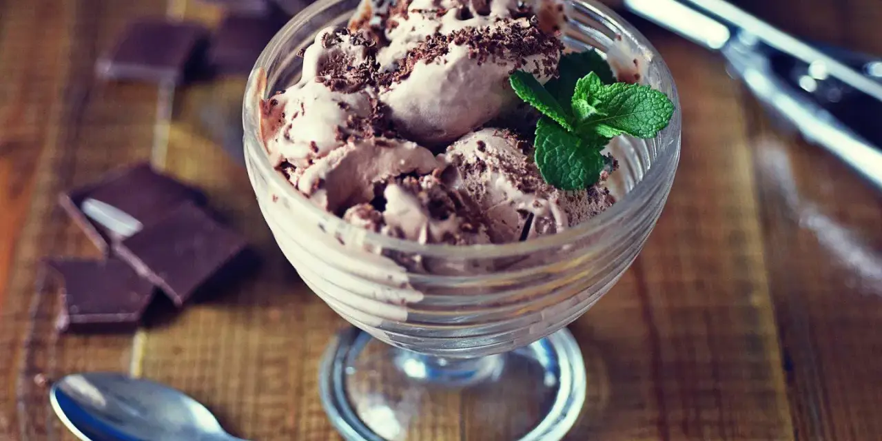 Pyszne lody nuii w pucharku, posypane czekoladą i udekorowane miętą. Obok leżą kawałki czekolady i łyżeczka.