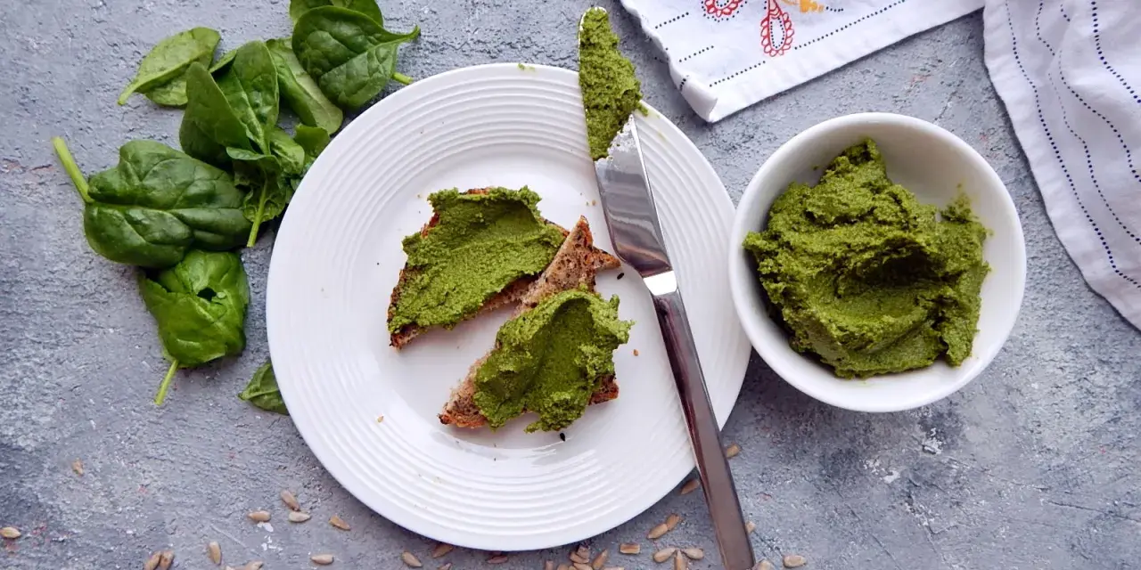 Pesto ze szpinaku do słoików na chlebie z pestkami słonecznika. Świeży szpinak obok talerza.