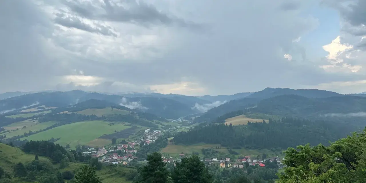 Malownicza panorama z widokiem na wieś w dolinie, otoczoną zielonymi wzgórzami i lasami. Nadciągają chmury, tworząc dramatyczne niebo nad lesnickie sedlo.