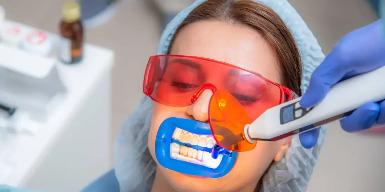 Kobieta przechodzi zabieg wybielania zębów u dentysty. Specjalista używa lampy aktywującej żel wybielający.