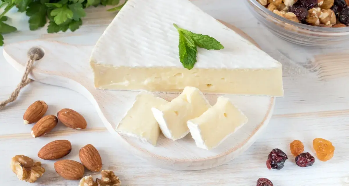 Queso brie en cuña y trozos, decorado con menta. Alrededor hay almendras, nueces y frutos secos. Ideal para quienes buscan información sobre queso brie calorias.