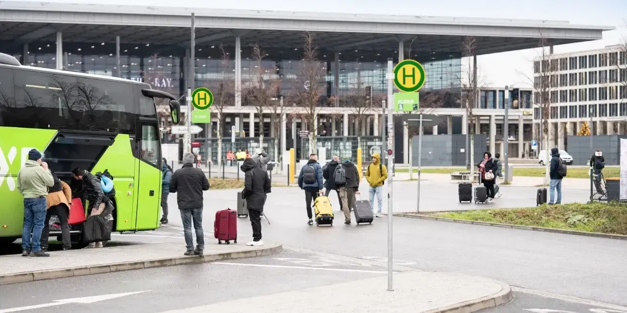 Ludzie z bagażami wsiadają do zielonego autokaru przed nowoczesnym budynkiem, przypominającym lotniska w Berlinie.
