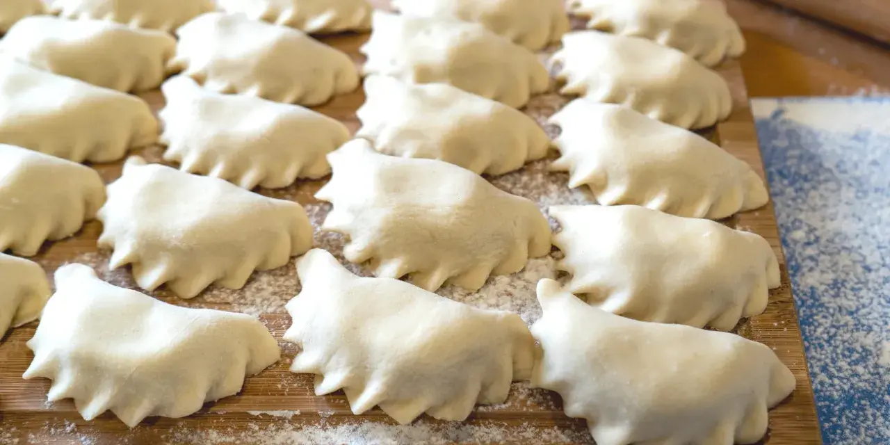 Surowe pierogi ułożone rzędami na drewnianej desce, posypanej mąką.