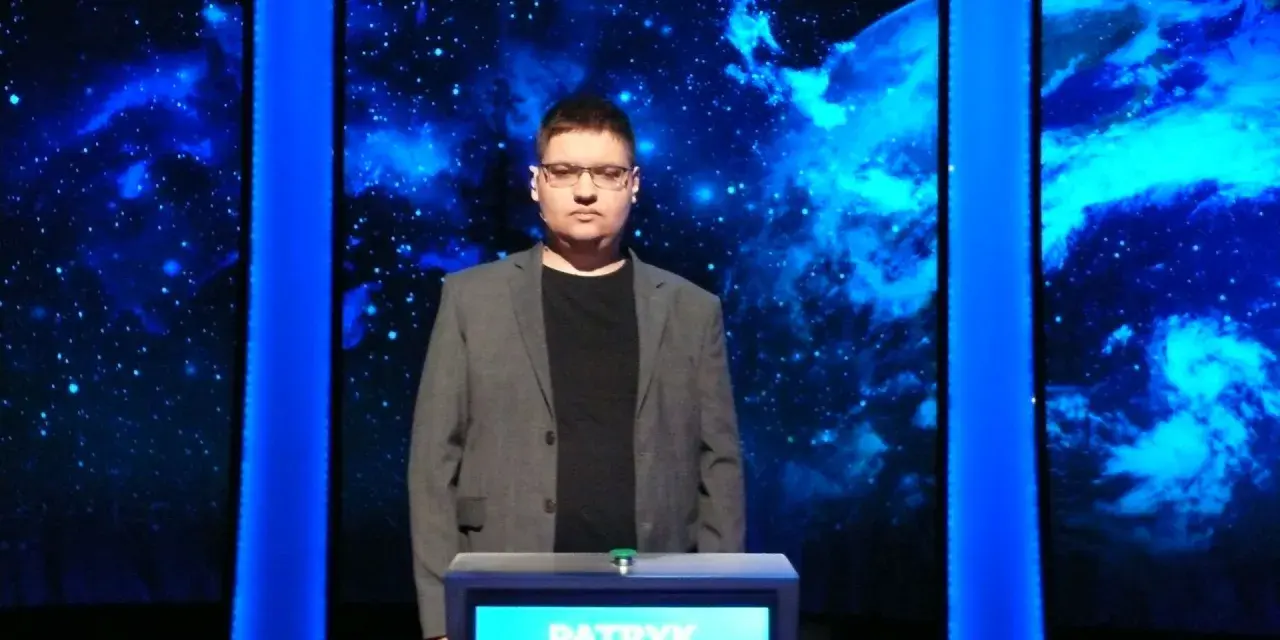 Patryk w szarym garniturze stoi przed niebieskim tłem z gwiazdami.