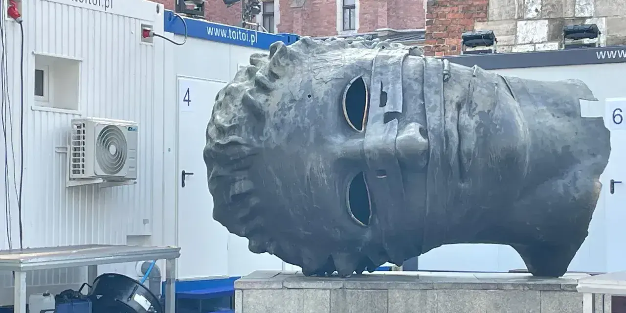 Monumentalna głowa na rynku w Krakowie, z pustymi oczodołami, stoi obok białych kontenerów.