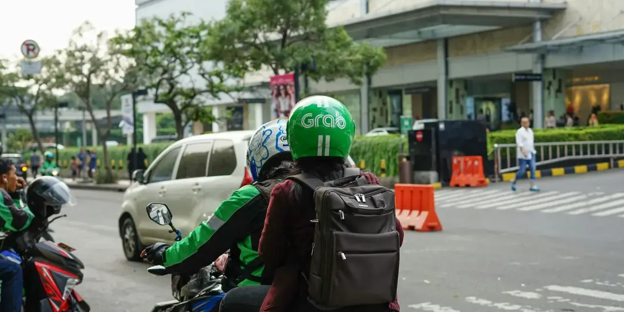 Zwei Personen auf einem Roller, der Fahrer trägt einen grünen Helm mit dem "Grab" Logo. Sie sind unterwegs in Thailand, um mit der Grab App Kunden zu transportieren.