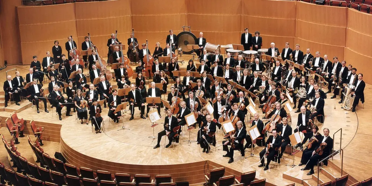 Ein großes Orchester mit vielen Musikern, die klassische Instrumente wie Violinen, Celli und Blasinstrumente spielen, posiert auf einer Bühne.