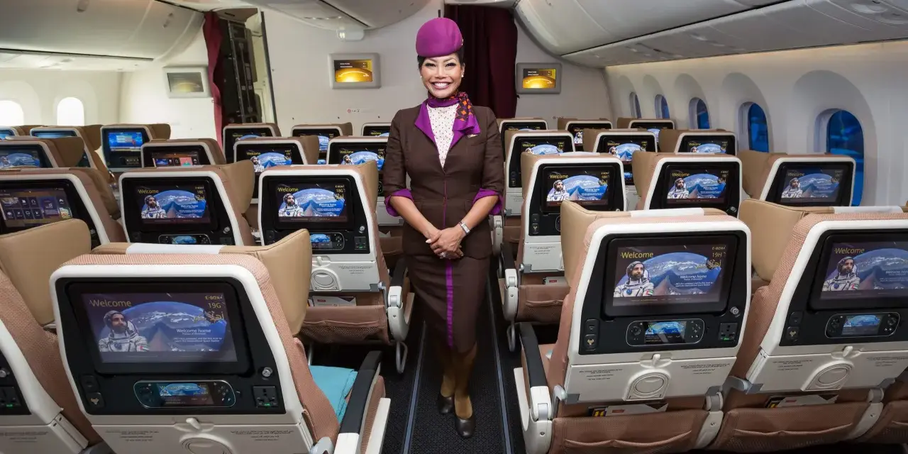 Uśmiechnięta stewardessa Etihad Airways w brązowym mundurku stoi w rzędzie foteli. Ekraniki pokazują astronautę. Dobre etihad airways opinie.