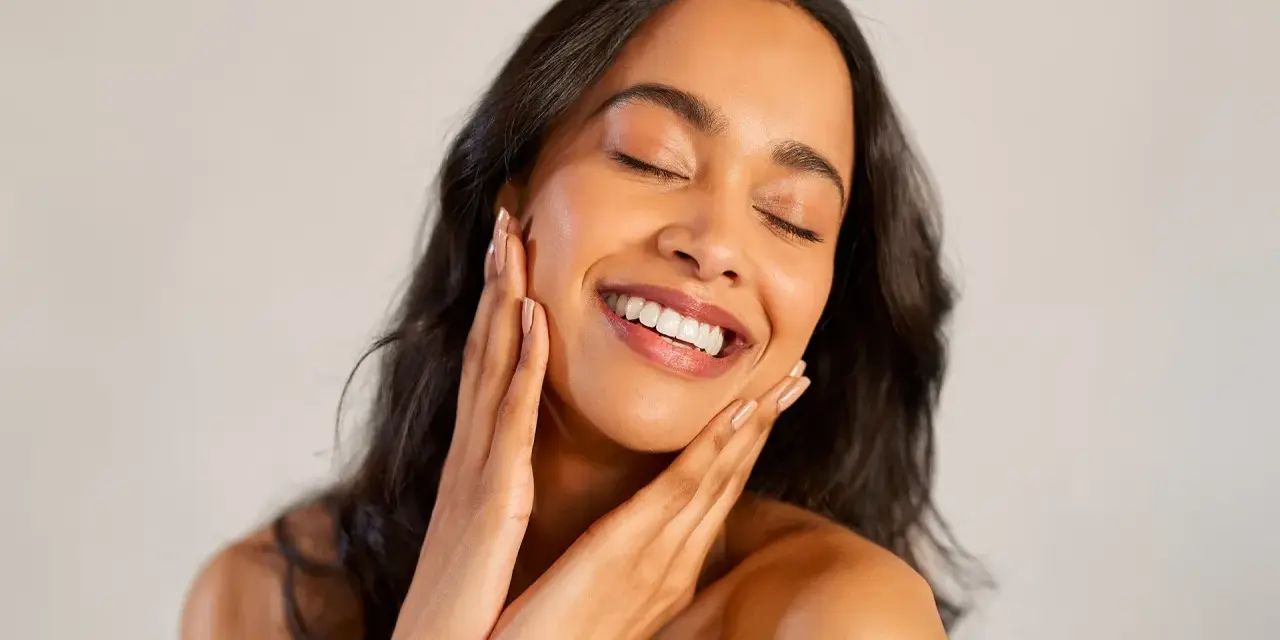 Une femme au sourire radieux caresse son visage, révélant une peau glowy éclatante de santé.