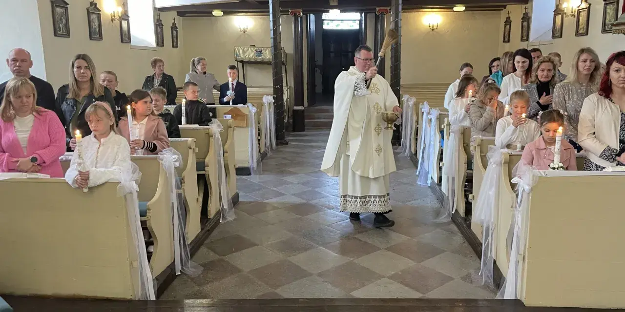 Uroczyste odnowienie przyrzeczeń chrzcielnych w kościele. Dzieci z zapalonymi świecami siedzą w ławkach, a ksiądz błogosławi zgromadzonych.