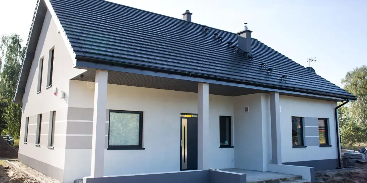 Nowy dom z szarą dachówką i jasną elewacją, ozdobioną subtelnymi, poziomymi pasami. Trwają prace przy malowaniu elewacji, tworząc nowoczesny wygląd.