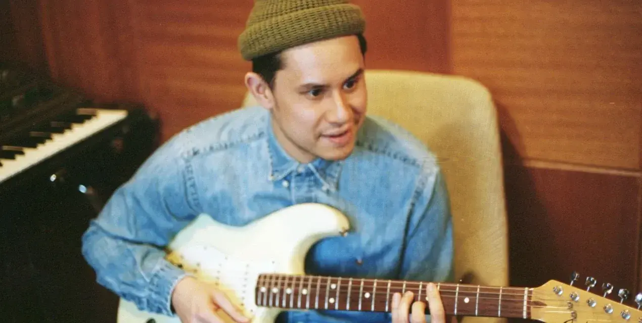 Frank Dukes w zielonej czapce i dżinsowej koszuli gra na gitarze Fender Stratocaster w studiu.