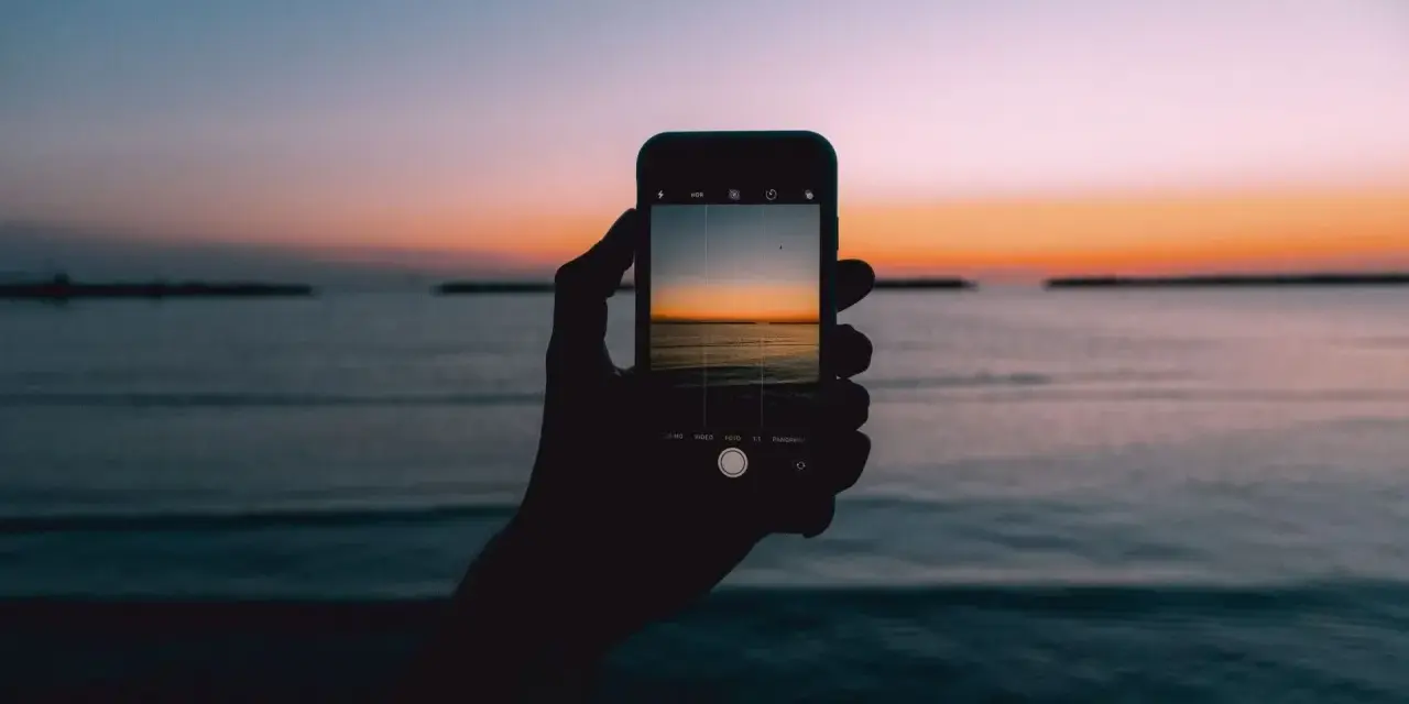 Dłoń trzyma smartfon, na ekranie którego widać piękny zachód słońca nad morzem. Idealny kadr, by nauczyć się, jak fotografować zachód słońca.