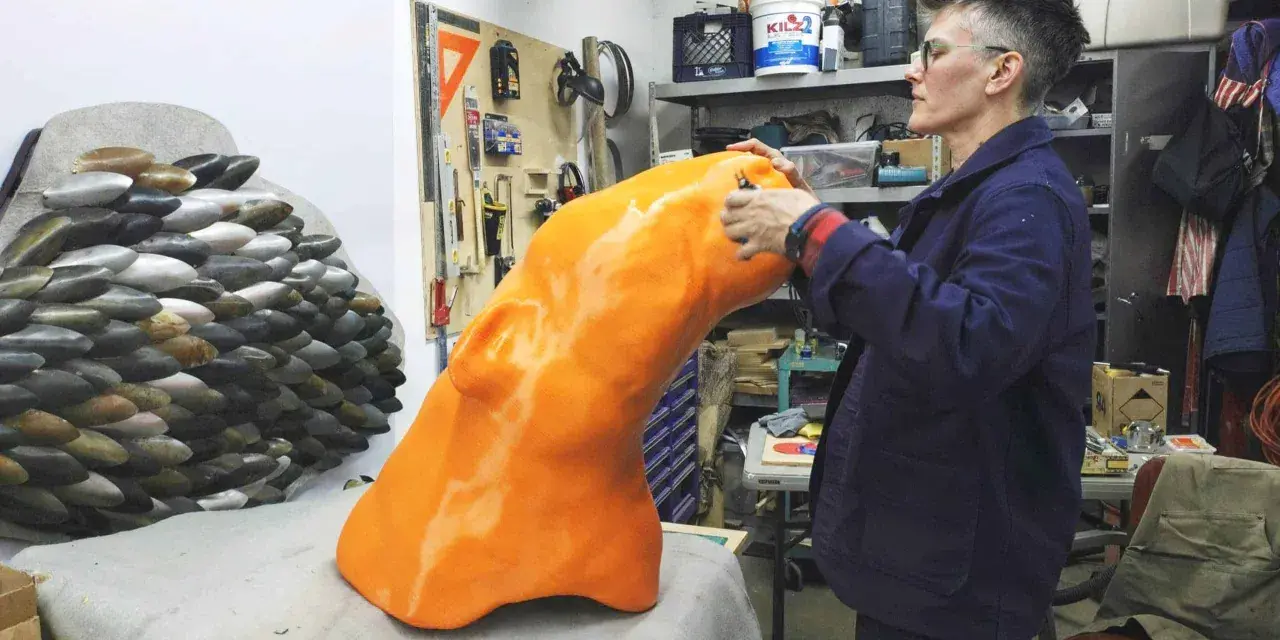 Artist applies finishing touches to a large, orange, 3D printed object, part of a project creating large objects.
