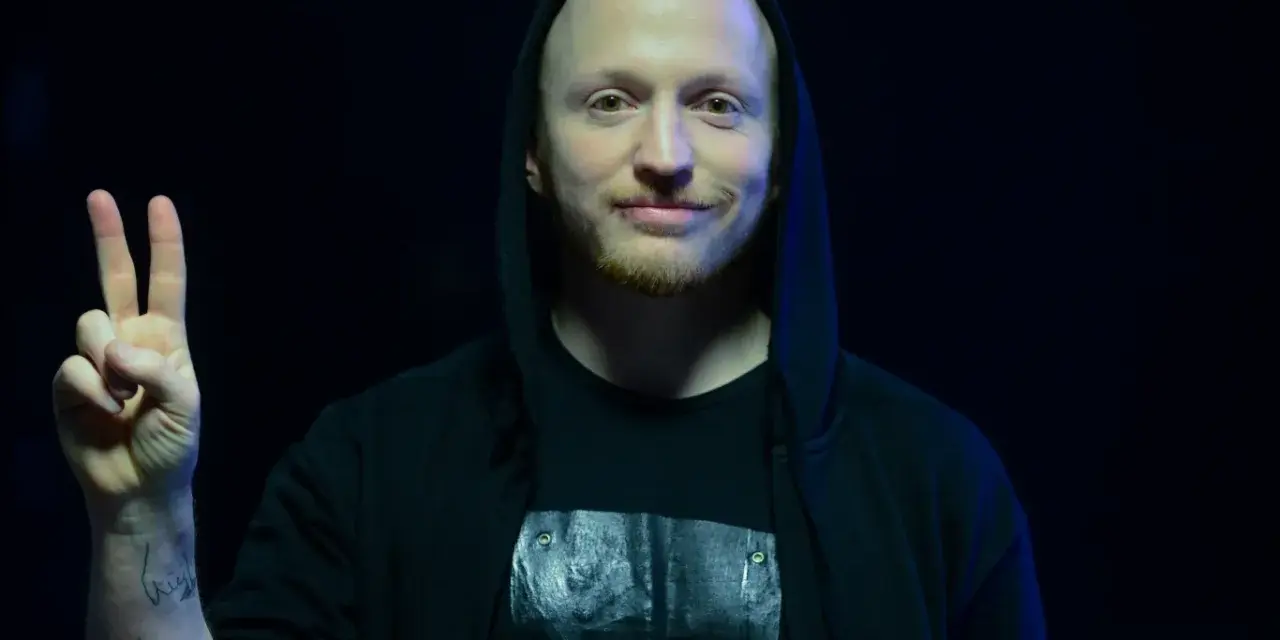 Wojtek Urbański w czarnej bluzie z kapturem pokazuje znak pokoju.
