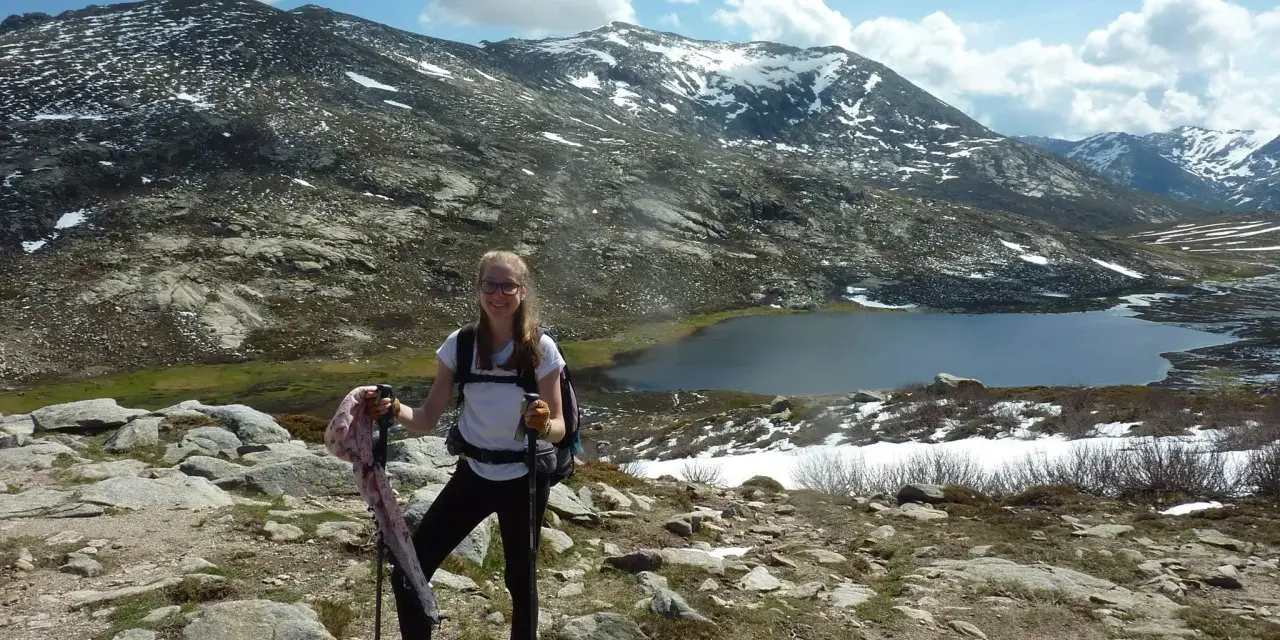 Jeune randonneuse souriante avec bâtons, face à une **mare** et des montagnes enneigées. La **difficulté** du sentier **nord** est palpable.