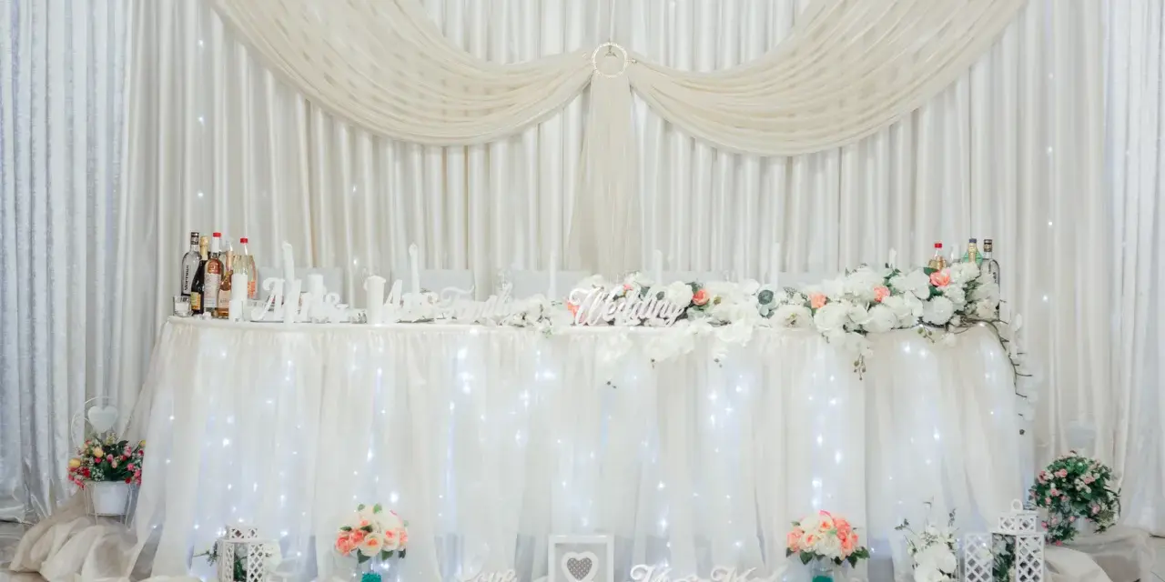 Elegancka ścianka za parą młodą inspiracje: białe zasłony, kwiaty, napisy "Wedding", "Love", "Mrs & Mr".
