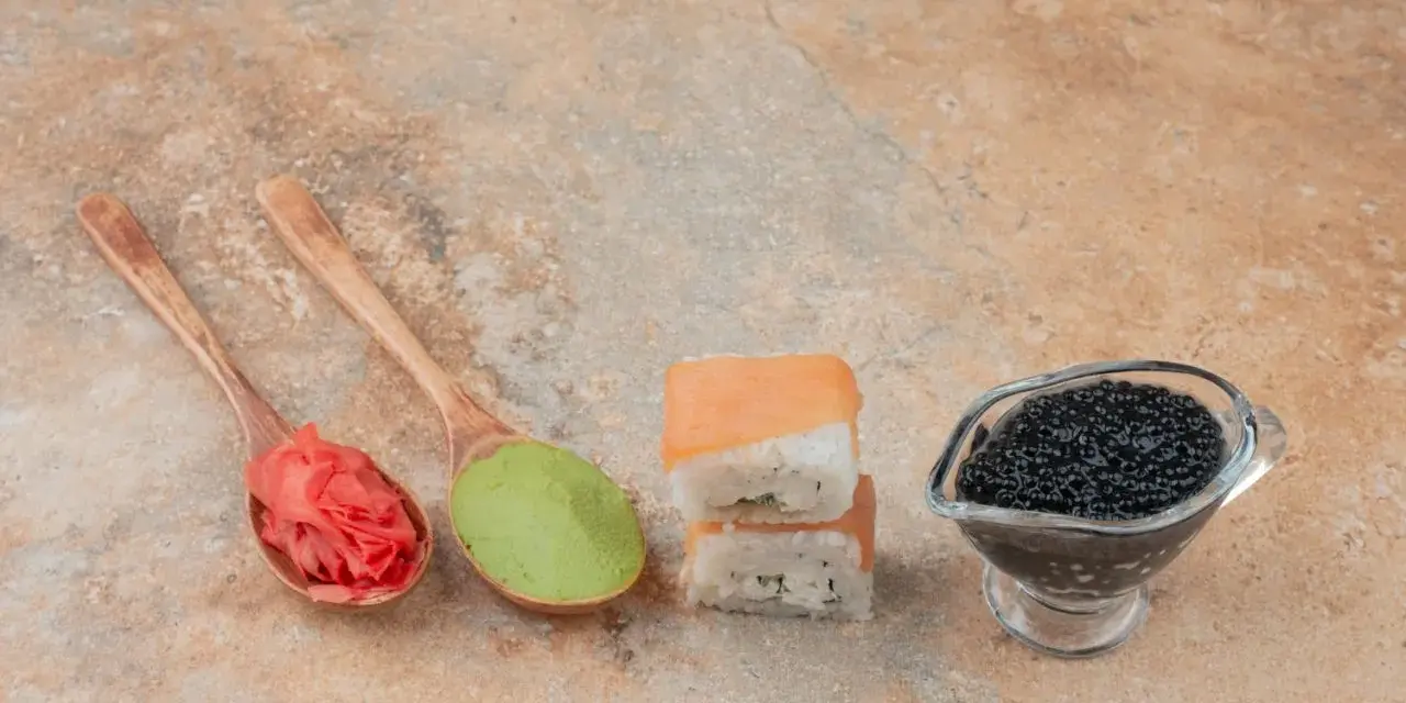 Na drewnianych łyżkach różowy imbir do sushi i zielone wasabi. Obok dwa kawałki sushi i sos sojowy z kawiorem.