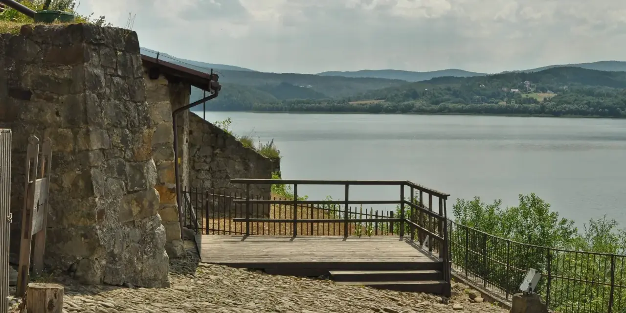 Kamienna wieża i drewniany taras z widokiem na spokojne jezioro dobczyckie. W oddali zalesione wzgórza.