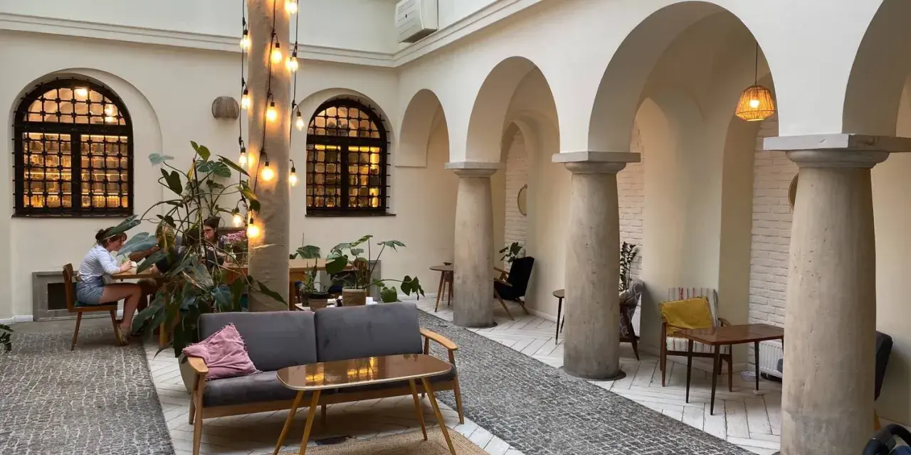 Przytulna kawiarnia patio z łukami i kolumnami. Goście siedzą przy stolikach, a obok stoi wygodna sofa. Widać rośliny i dekoracyjne lampki. Menu czeka na odkrycie.